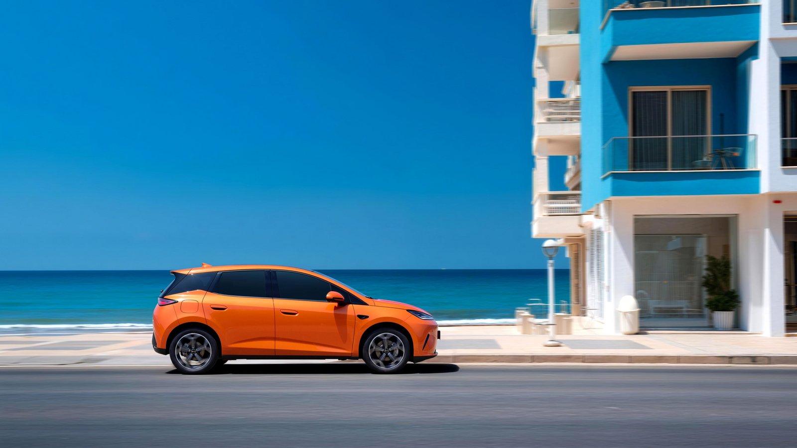 Seitliche Ansicht eines orangefarbenen BYD Dolphin G DM-i vor einem weißen Gebäude am Meer